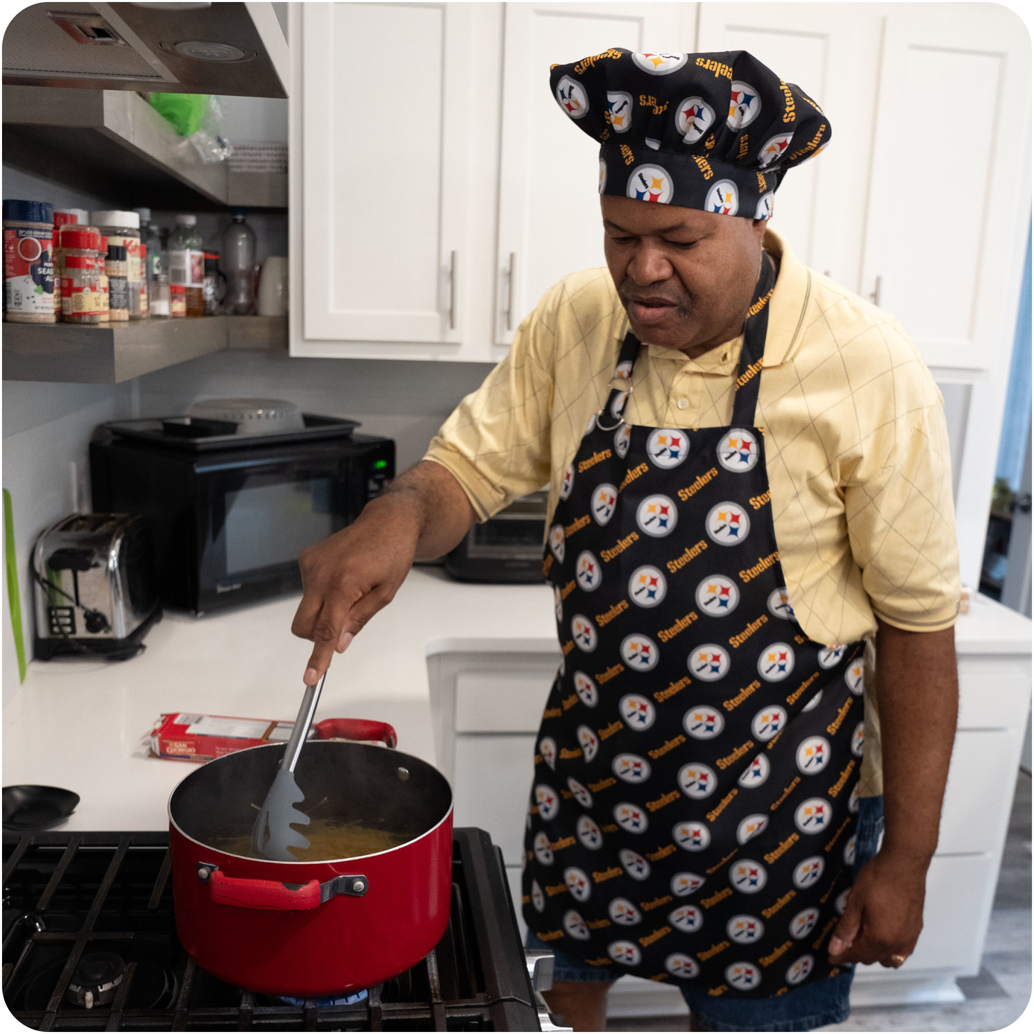 A man boiling pasta in his kitchen wearing a Steelers apron and chef hat