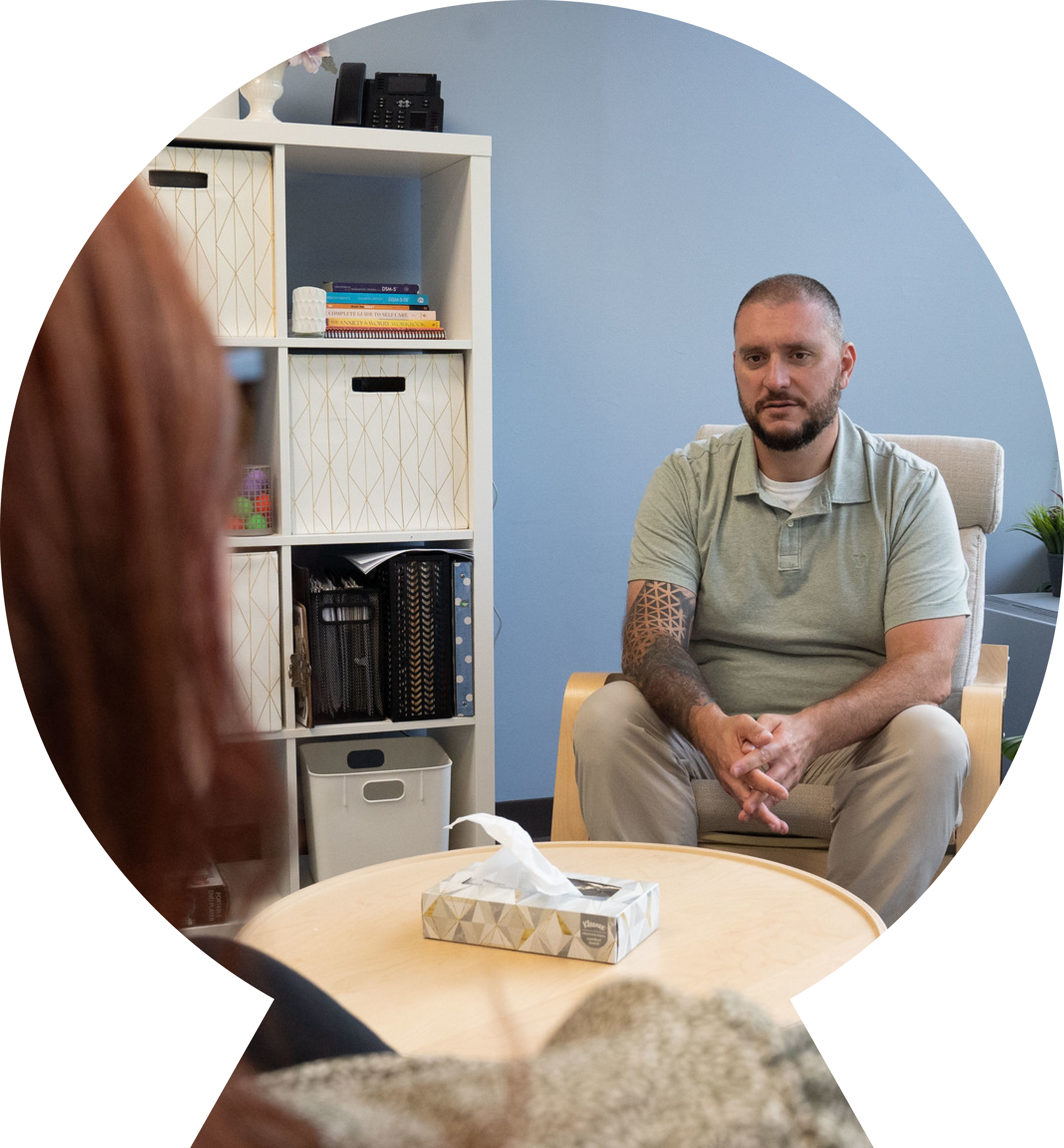 Young white man with tattoos sitting on a chair in an office talking to a red-haired woman.