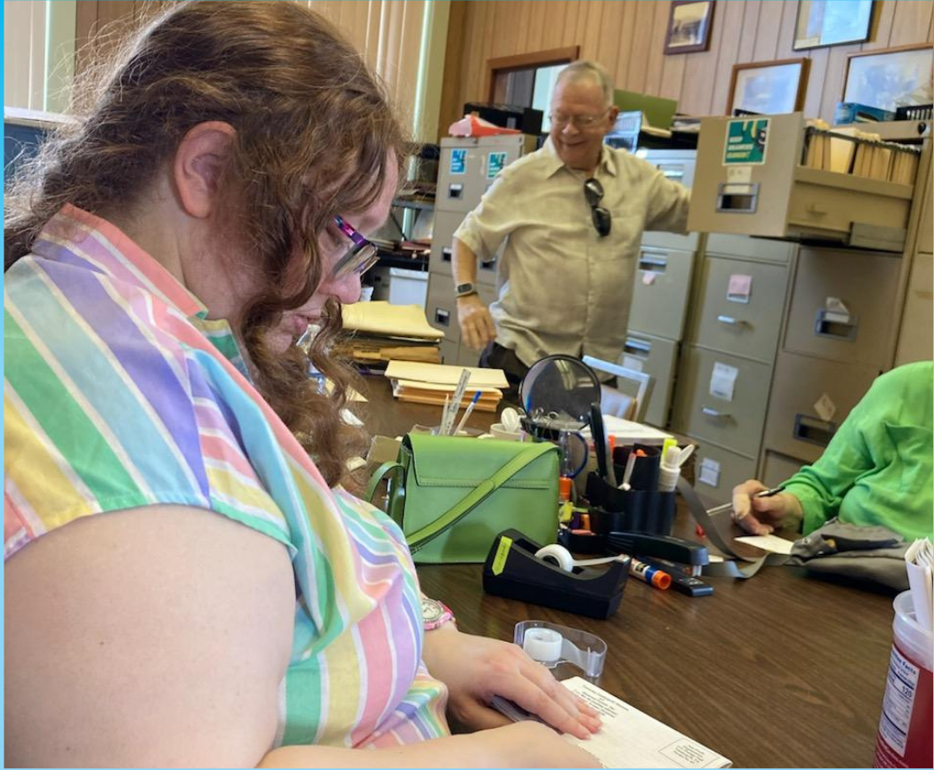 Young woman in the foreground of the photo in a rainbow striped shirt working. Older man in the background looking at a file drawer.