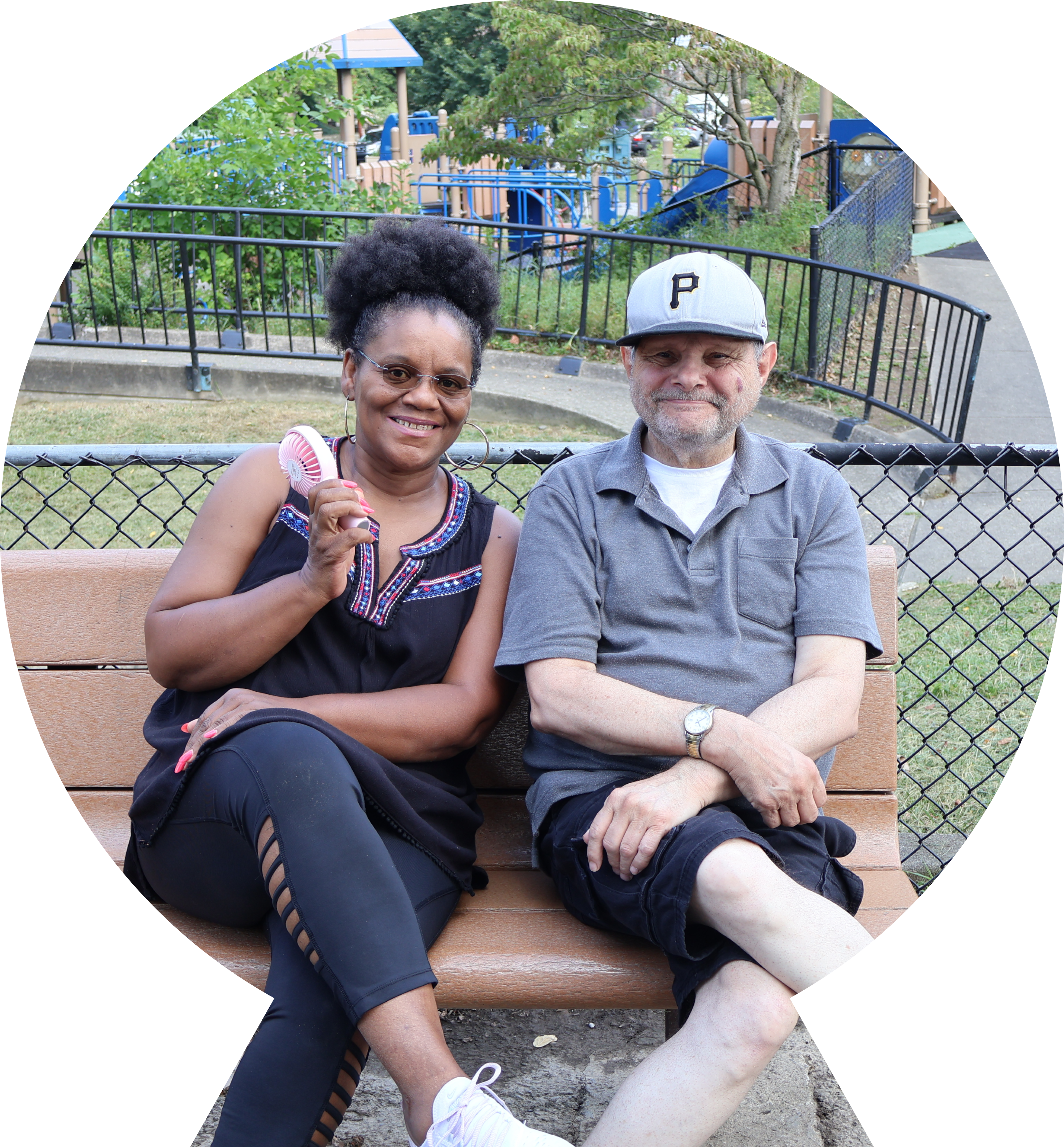 Woman with fan and man in a ball cap sit on a park bench smiling for the camera